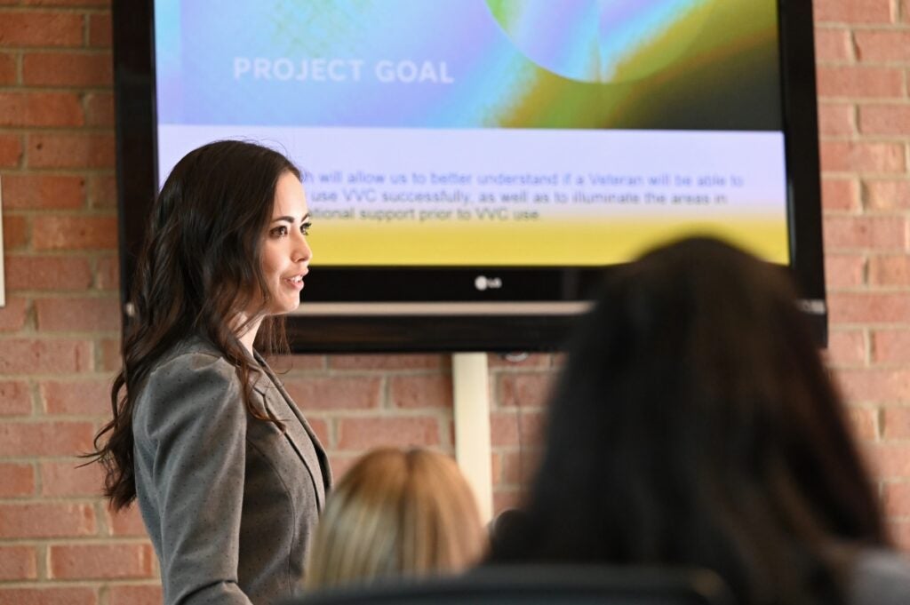 A woman with long dark hair in a gray suit jacket stands in front of a presentation screen.