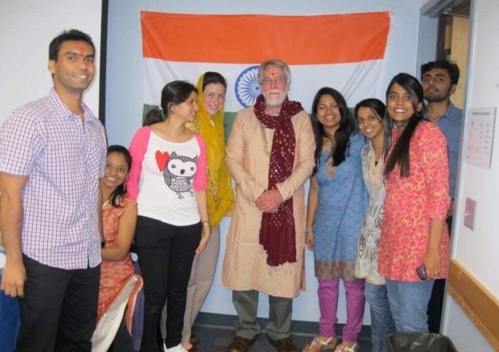 Students from India stand next to a man with a white beard and hair wearing a traditional yellow Indian coat and long burgandy scarf