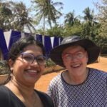 Woman in blue glasses smiling at camera with another woman smiling wearing a black rim hat and red sunglasses