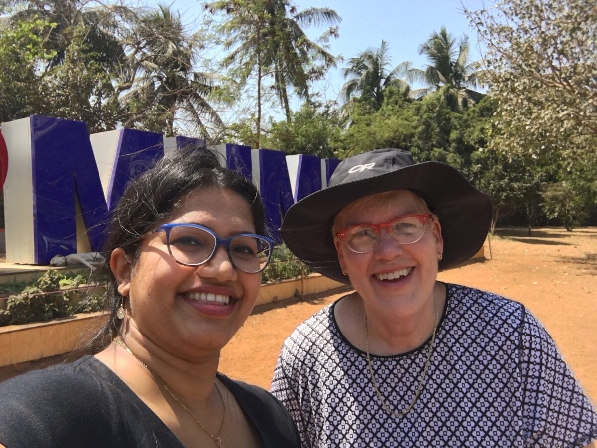 Woman in blue glasses smiling at camera with another woman smiling wearing a black rim hat and red sunglasses