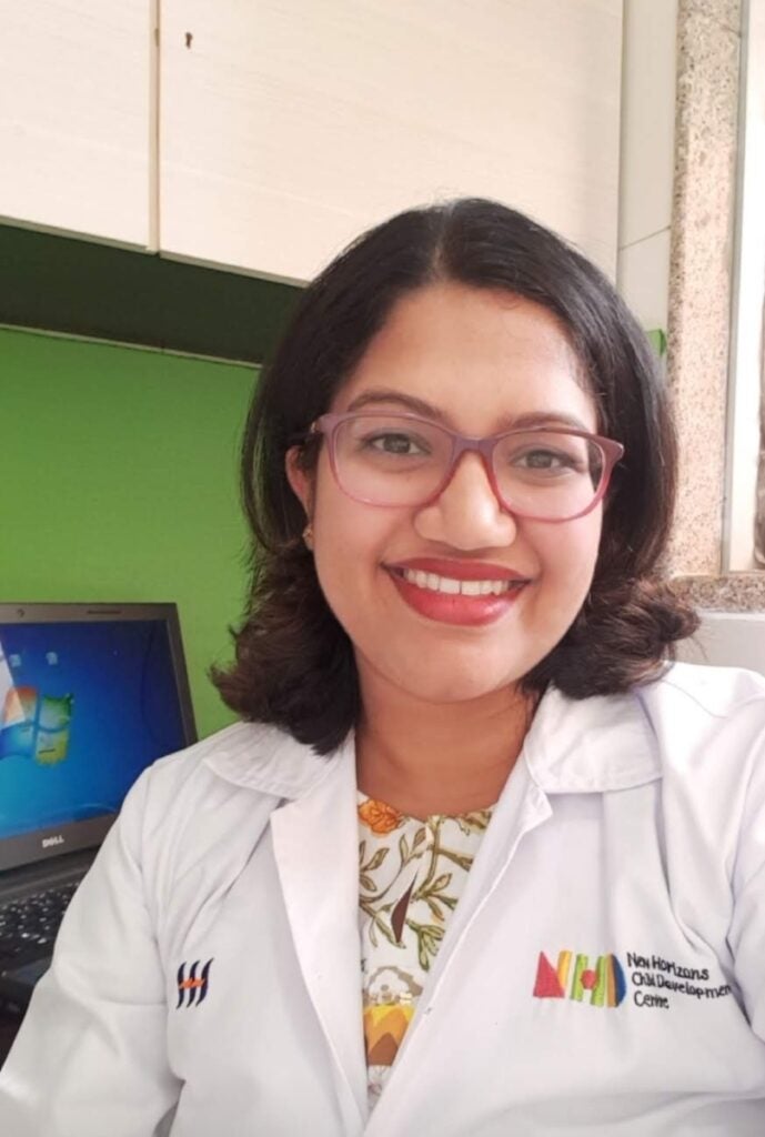 Woman with medium length brown hair with red glasses wears a white lab coat smiling at the camera