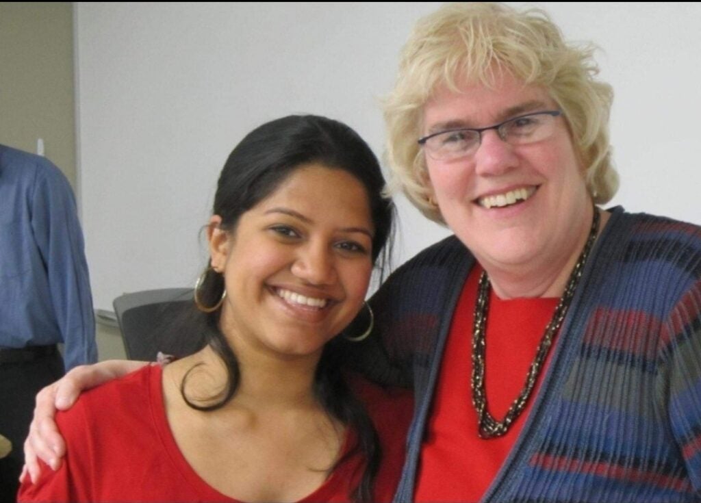 A woman with long dark hair in a ponytail and gold hoop earrings smiles at the camera with a woman with short blond hair and glasses