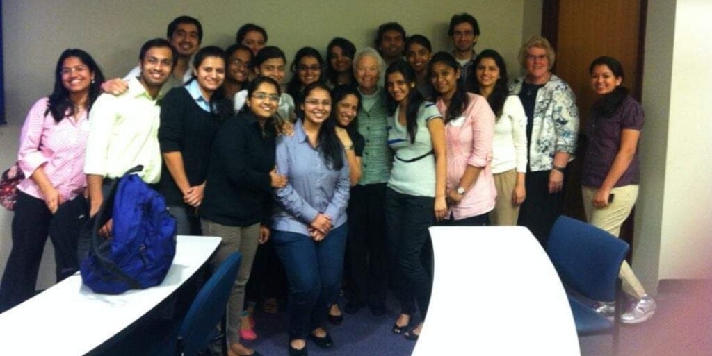 A classroom of students stands next to a female professor in a green sweater in the center