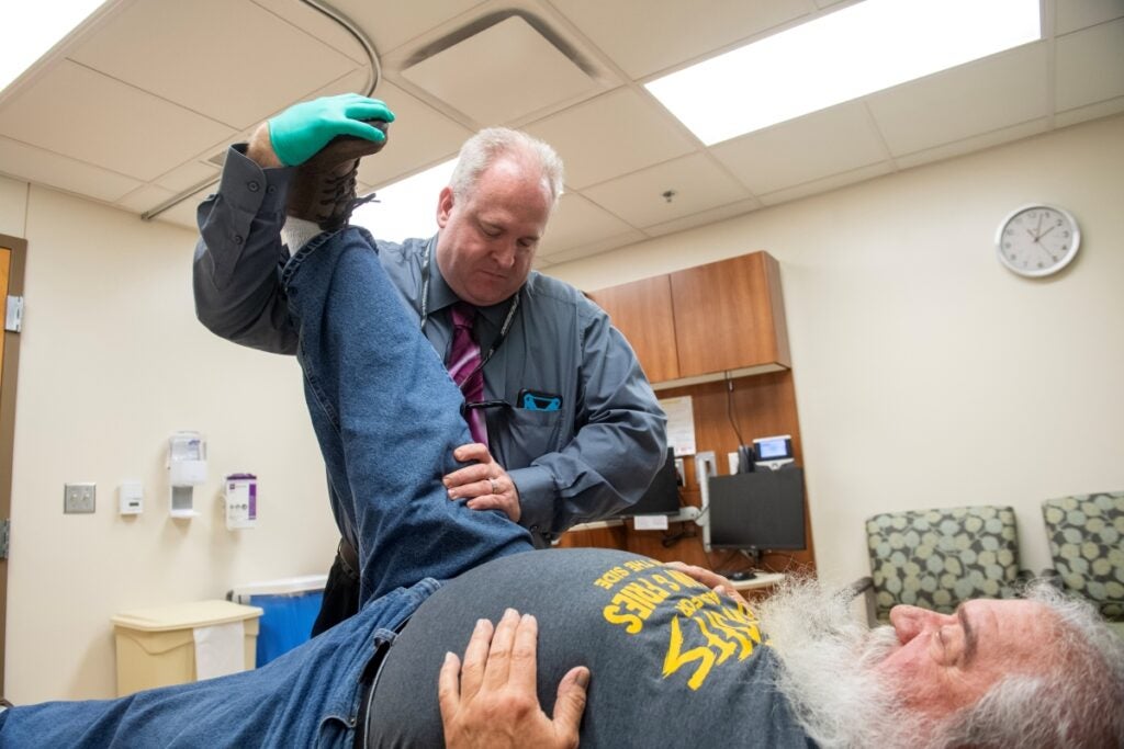 Faculty member stands over a patient laying on a table and holds their right leg up to demonstrate stretching.