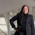 A woman with dark skin and long braided hair in a black suit poses for a photo