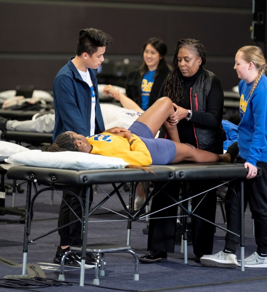 A woman with dark skin and long dark hair bends a patient's leg on a table while two students look on.