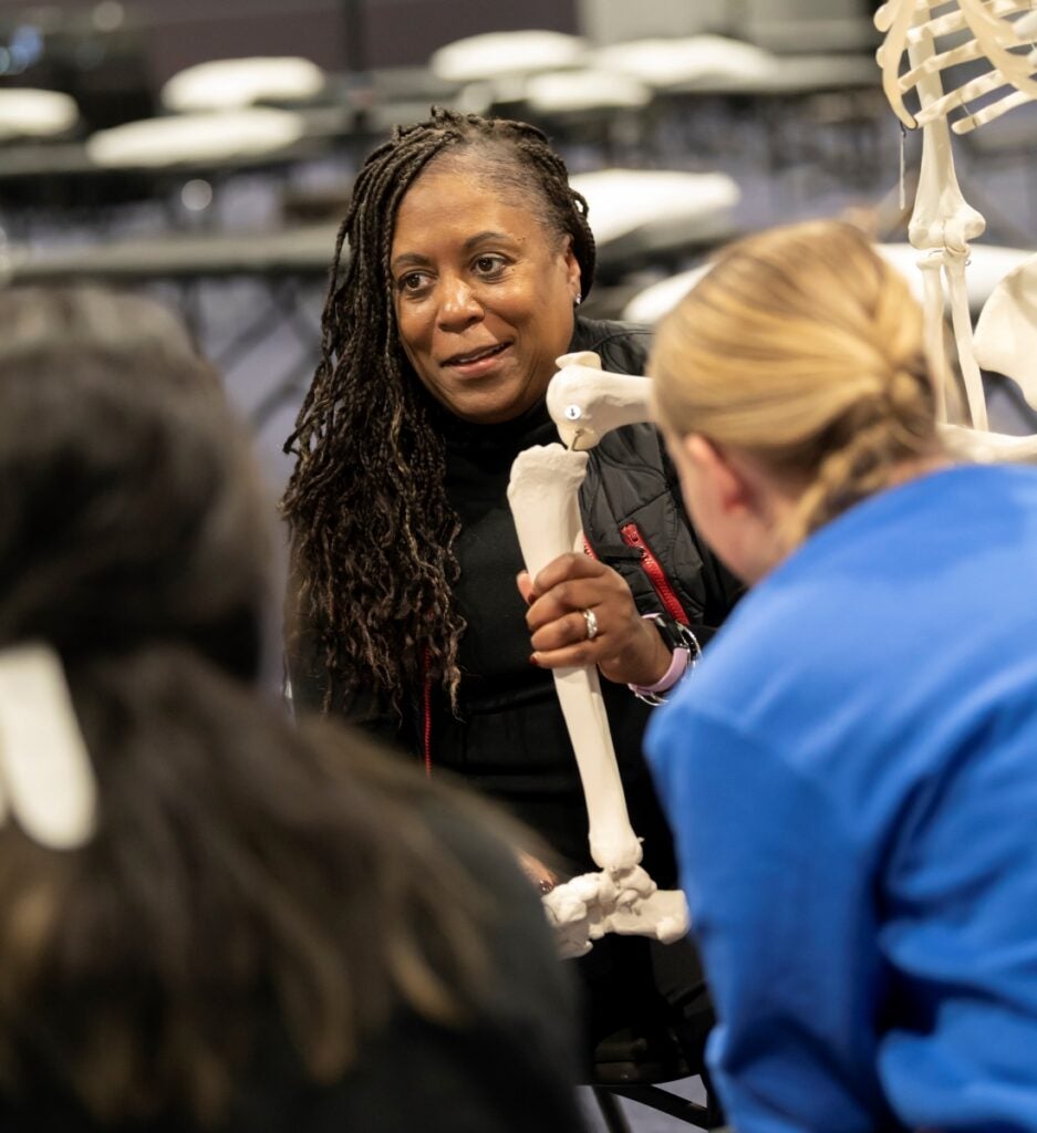 A woman with dark skin and long dark braids holds a skeleton figure's leg for a demonstration to students