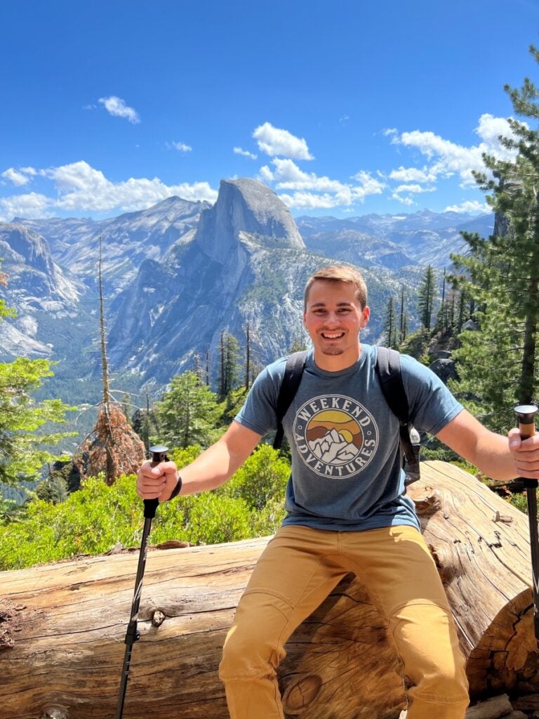 A man holding hiking poles sits on a tree log with the vista of Half Dome rock formation in the background