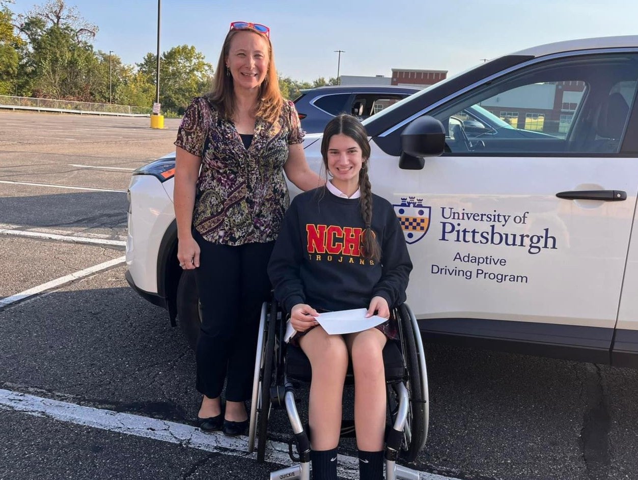 A woman with auburn hair in a brown patterned blouse standing next to a woman in a wheelchair who has her brown hair in a braid and is wearing a navy blue graphic sweatshirt.