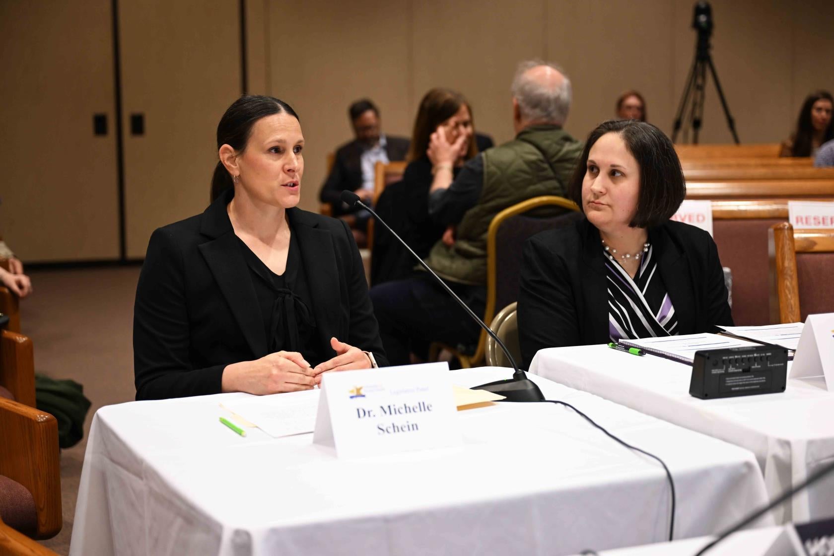 Woman with black hair wearing a black blazer sits at a table covered in white tablecloth while speaking into a microphone. Next to her, a woman with short brown hair and a black and white striped blouse looks at her.