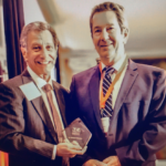 Two men in suits hold a small crystal award at a conference