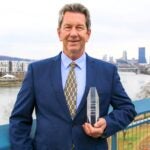 Man wearing a dark blue suit and yellow tie, standing on a balcony in front of the Pittsburgh skyline holding a crystal statue award in his left hand.