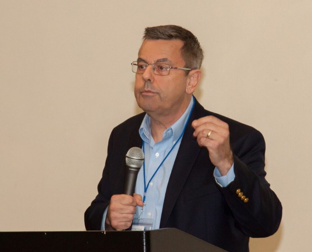 A man with short brown hair wearing a black suit jacket over a blue collared shirt while holding a microphone at a podium.