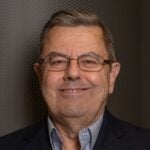 Man with brown hair wearing a black suit jacket over a gray collared shirt and wearing wired glasses.