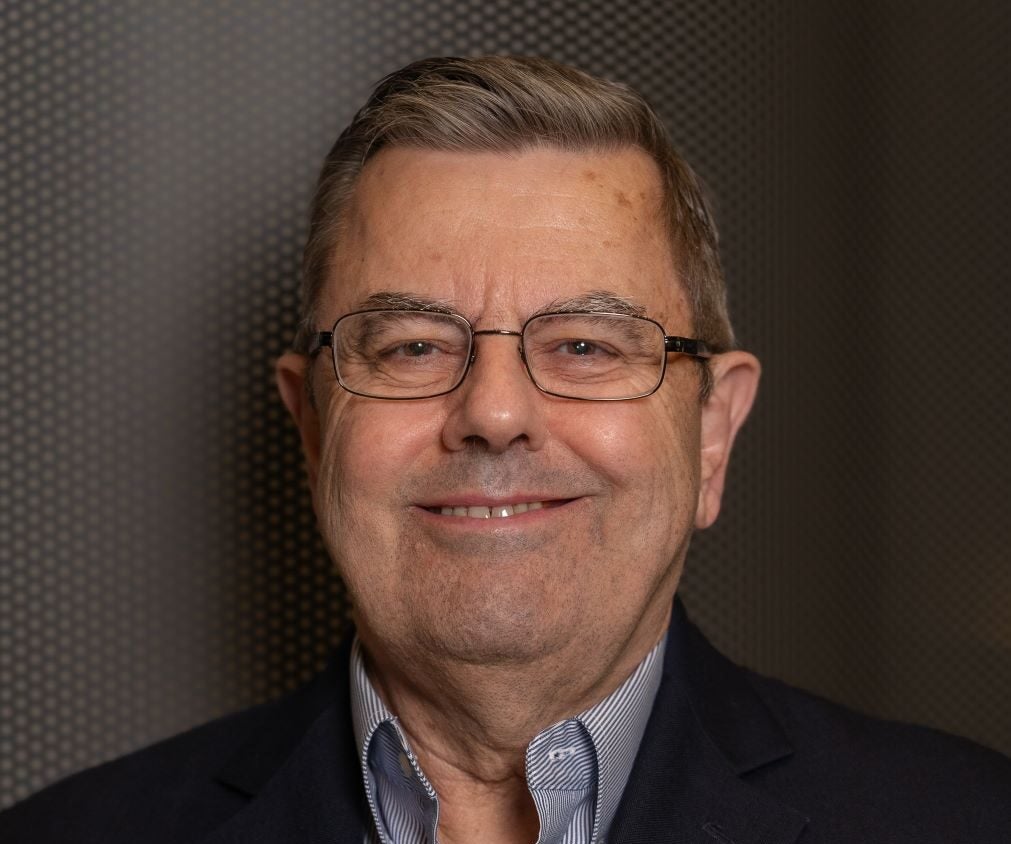 Man with brown hair wearing a black suit jacket over a gray collared shirt and wearing wired glasses.