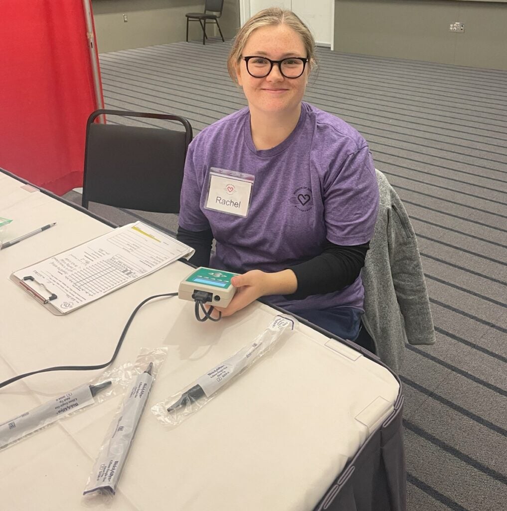 A student with blonde hair and glasses wearing a purple t-shirt sits at a table holding a device