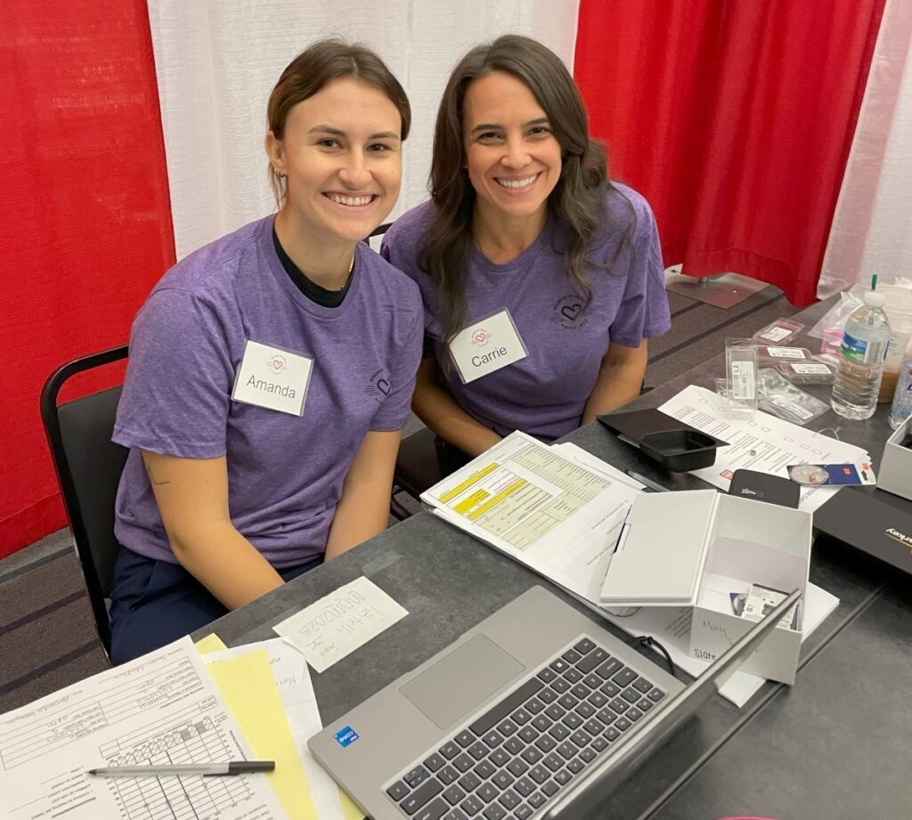 Two women wearing purple t-shirts sit at a table with a laptop computer and paperwork