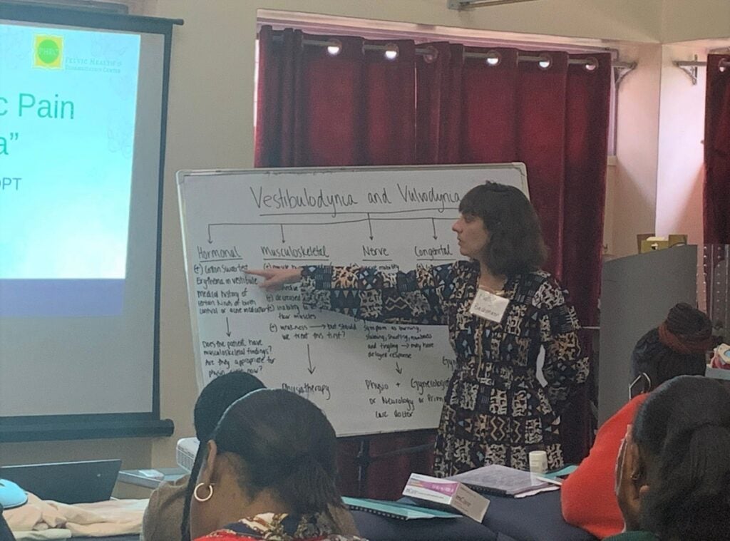 A woman with brown hair and long colorful dress points her right hand to a dry erase board with information written on it as she teaches a classroom