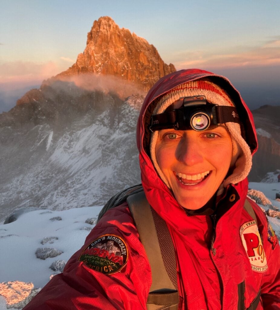 A woman in heavy mountaineering clothing and a headlamp smiles in a selfie while standing on top of a mountain at sunrise