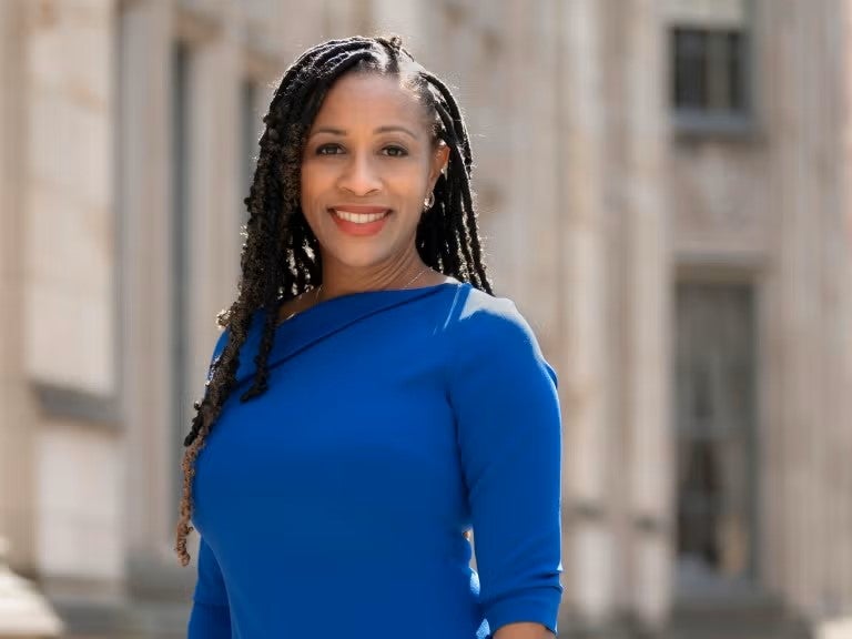 A woman with medium brown skin and long braided hair wears a bright blue dress