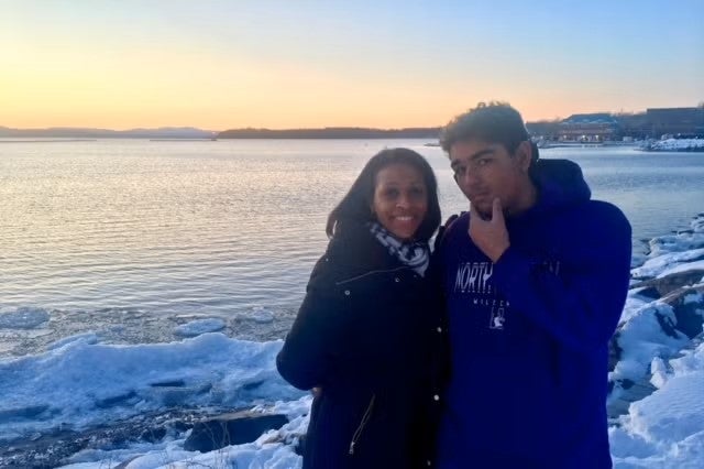 The sun sets along a lake with snow on the shore and a woman with medium brown skin and long black hair stands next to a young man with medium brown skin and short dark hair