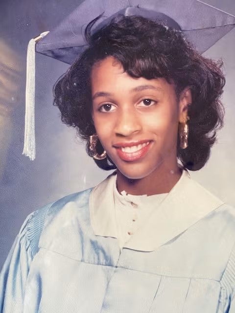 A young woman with medium brown skin and short black hair wears a light blue graduation cap and gown