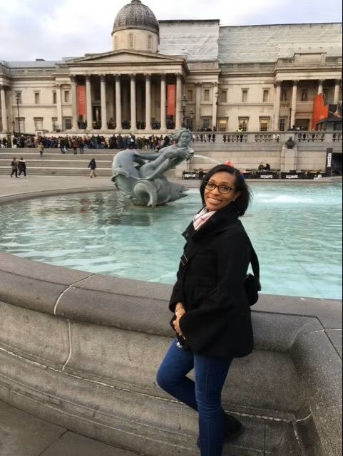 A woman with medium brown skin and short black hair wearing glasses poses in front of a fountain