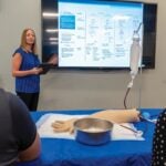 A woman with long blonde hair wearing a blue shirt stands at a presentation screen in front of a class