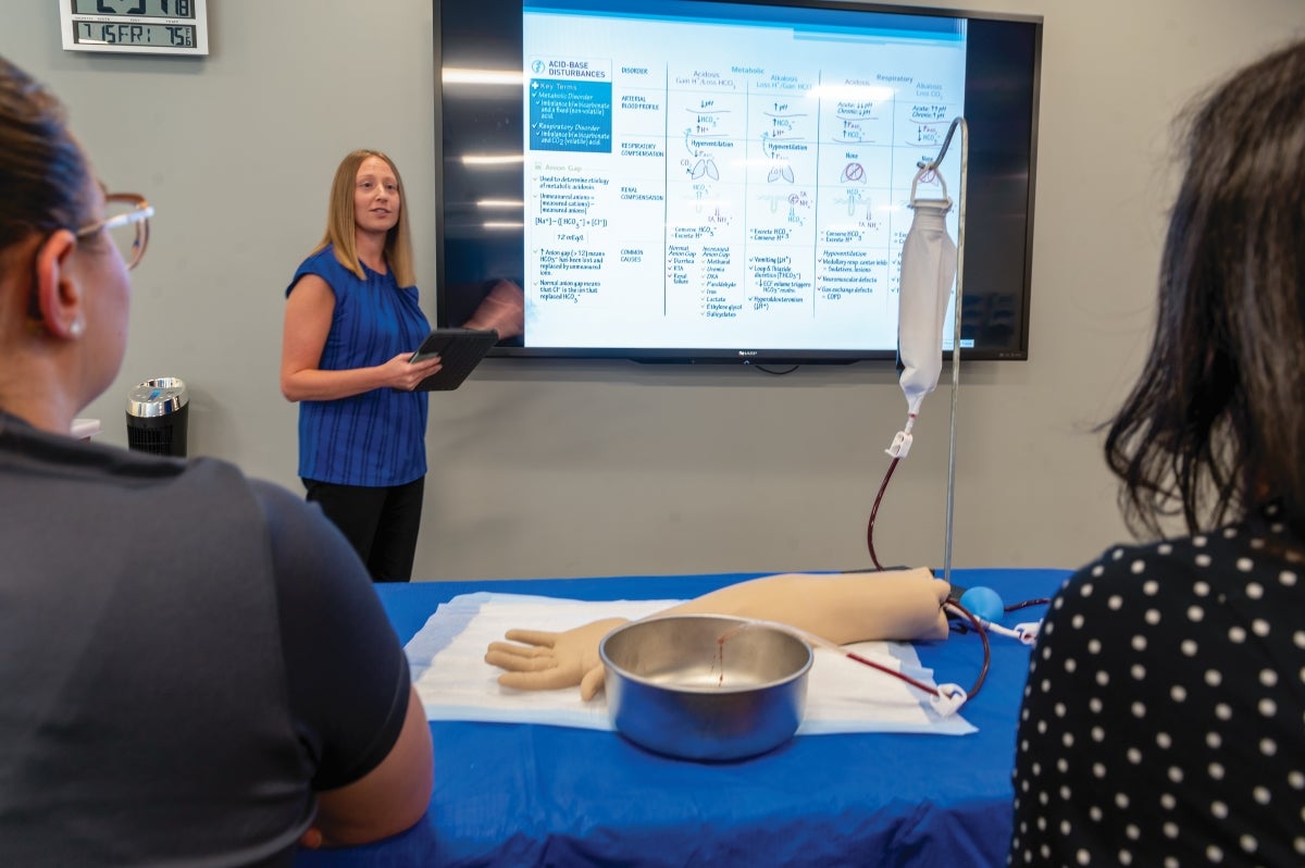 A woman with long blonde hair wearing a blue shirt stands at a presentation screen in front of a class