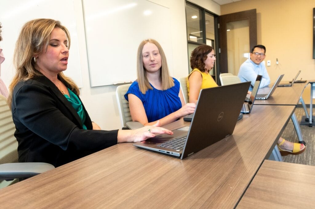 Four people sit at a long classroom table discussing information on a laptop computer.