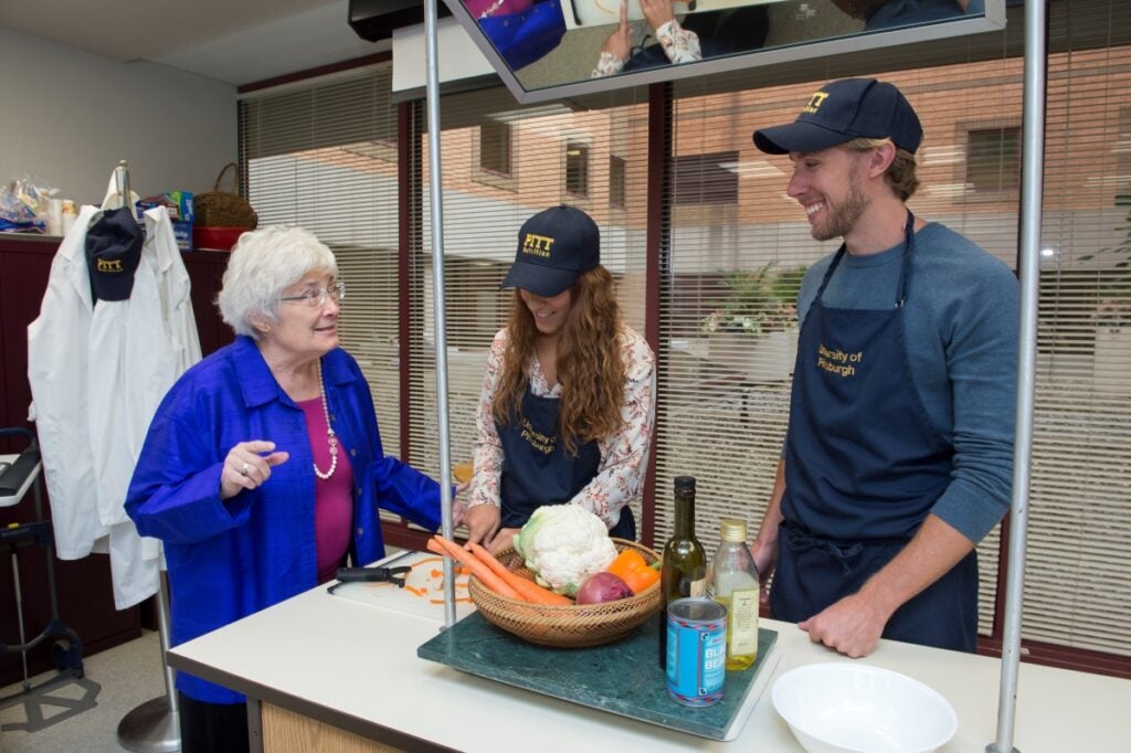 A teacher in a bright blue coat instructs two students cutting vegetables