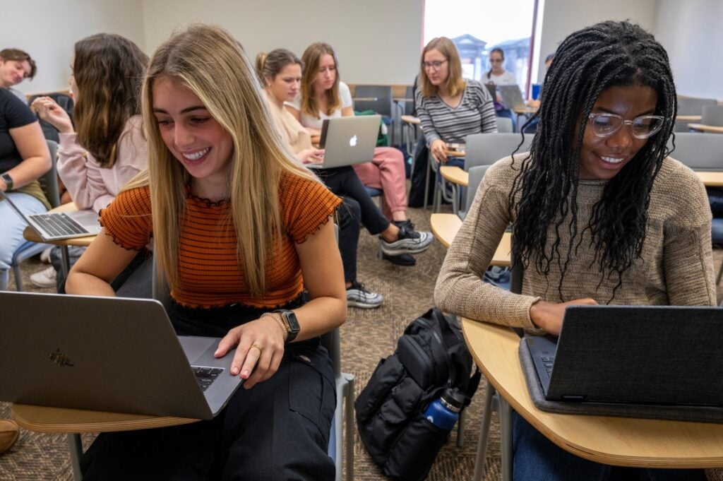 Two students sit in a classroom working at their laptops