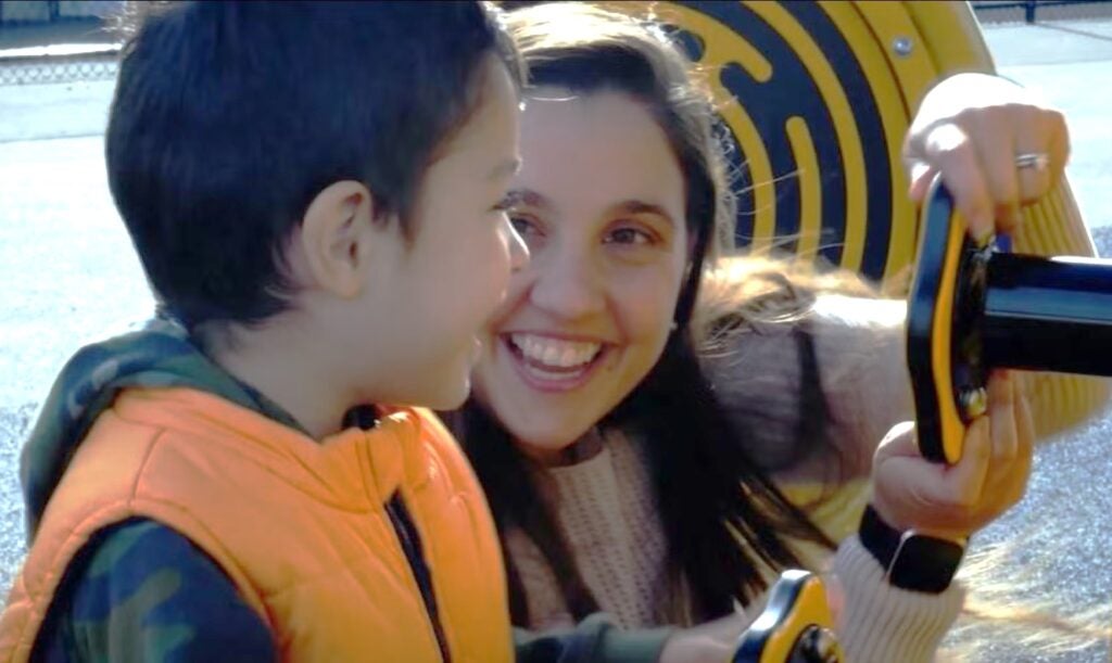 A woman with long brown hair holds a toy steering wheel while smiling at a small child.