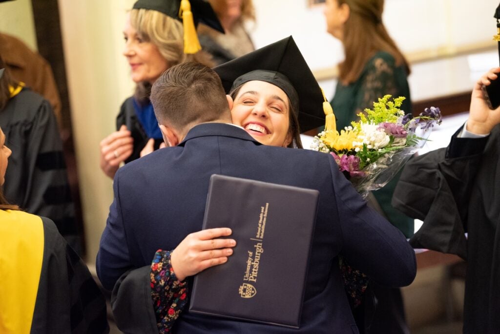 A woman in graduation regalia hugs a man and smiles