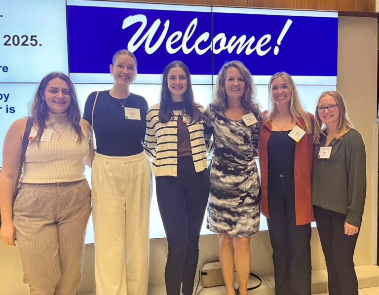 A group of six women wearing conference badges pose for the camera in front of a digital welcome sign