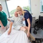 A woman with long blond hair helps an elderly man in a hospital gown to swing his legs onto a hospital bed