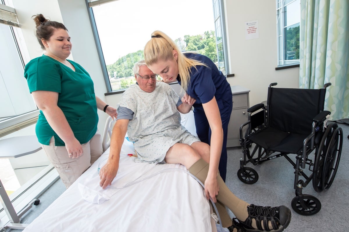 A woman with long blond hair helps an elderly man in a hospital gown to swing his legs onto a hospital bed