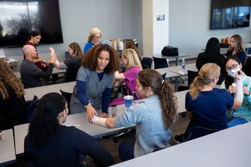 A woman with light brown skin and long brown curly hair stands and reaches out to a student with a bandage on their left arm