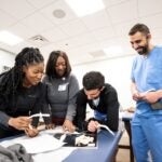 A group of four students look over a textbook while holding models of bone pieces