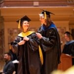 Two women wearing graduation regalia stand on a stage facing each other