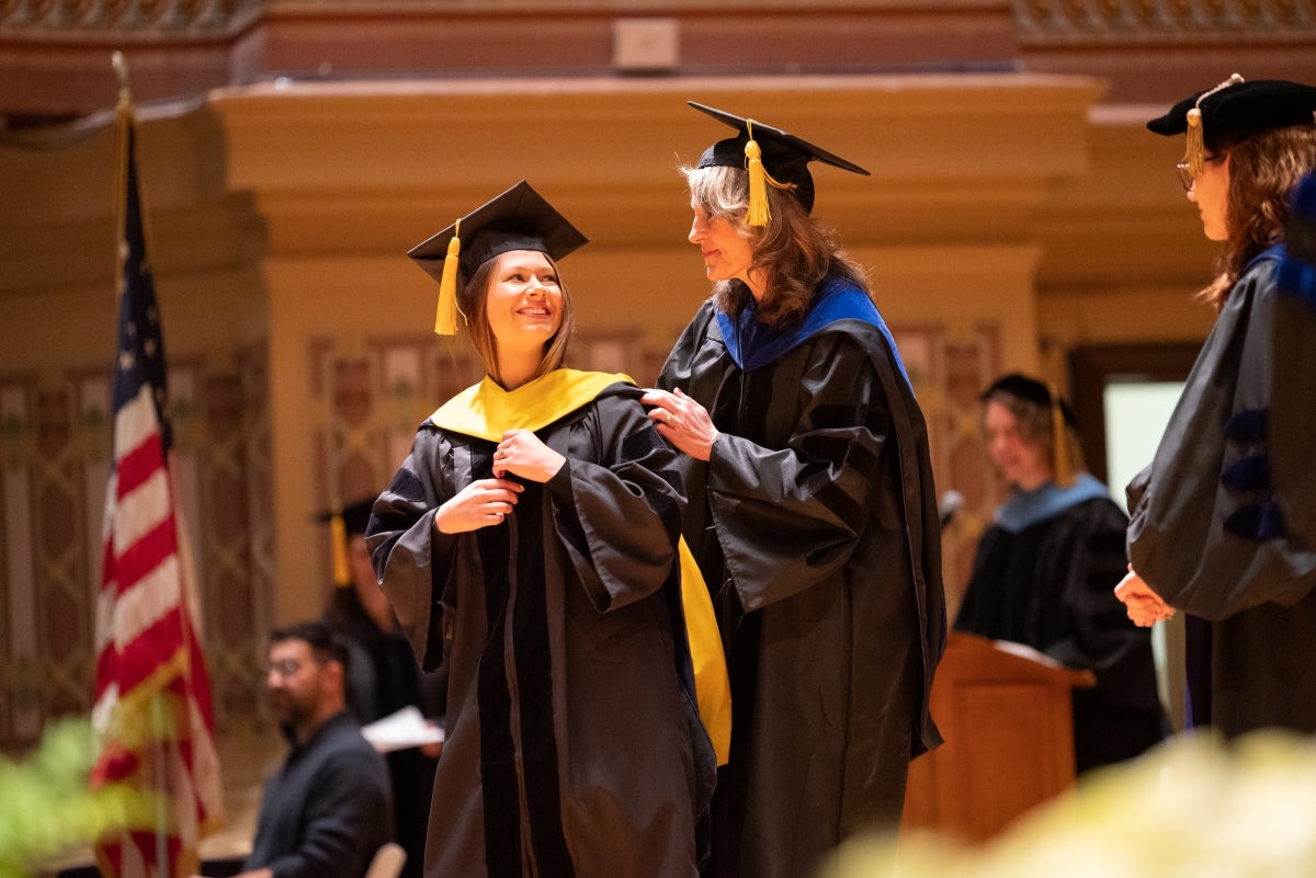 Two women wearing graduation regalia stand on a stage facing each other