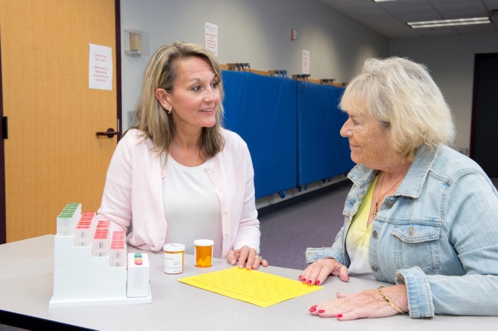 Two women sit at a table facing each other with bottles of prescription medication in front of them