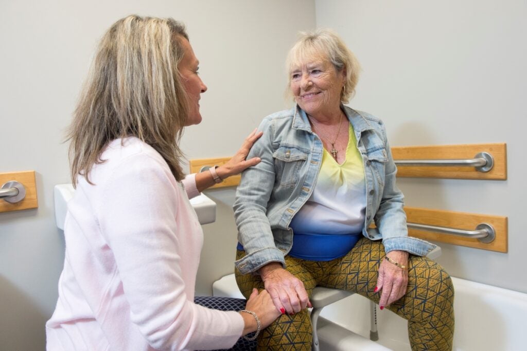 A woman in a pink sweater places her hand on the shoulder of an older woman sitting on a stool