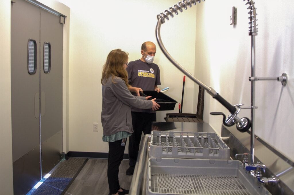 A woman with long blond hair helps a man with no hair to lift a bin to a washing station