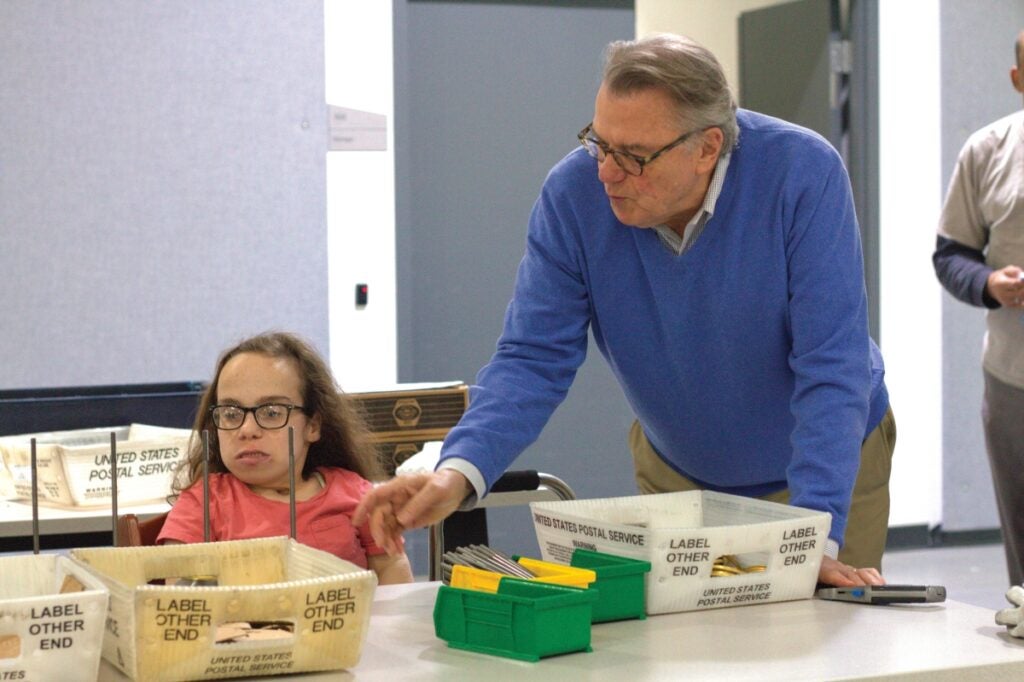 A man with gray hair and glasses wearing a blue sweater helps a woman sitting at a table