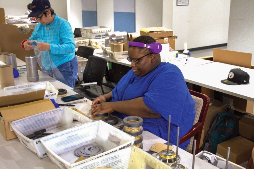 A woman with dark skin and short dark hair wearing a bright blue blouse sits at a table while working on circular equipment