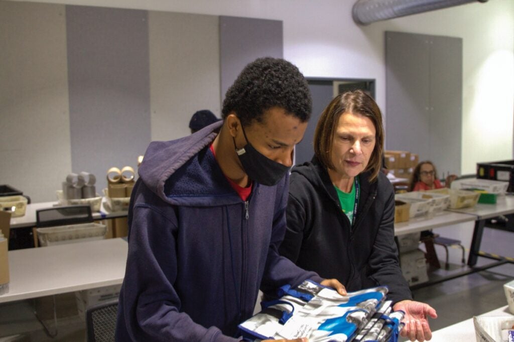 A woman with short brown hair helps a young man with medium brown skin and short dark hair to lift a pile of bags