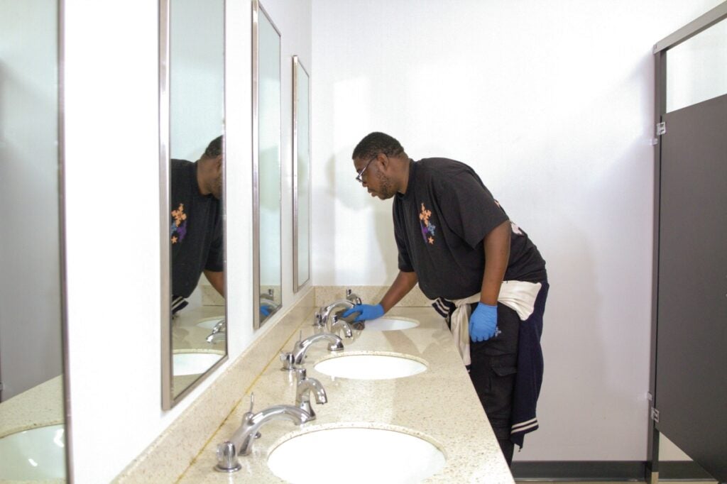 A tall man with brown skin and short dark hair and a beard cleans the sink in a bathroom