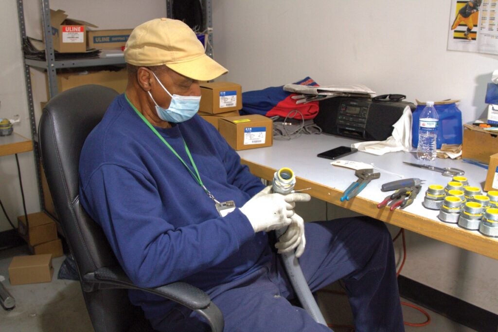 A man wearing a yellow baseball hat and mask sits in a chair and looks at a hose with a large attachment at its end