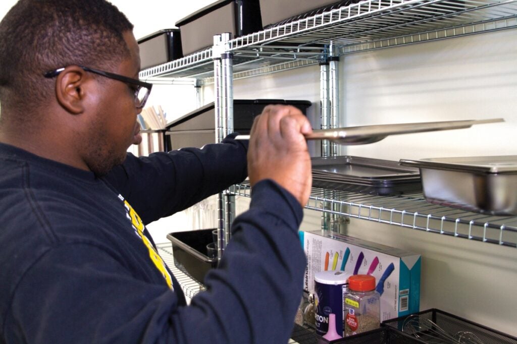 A man with dark skin and short dark hair wearing glasses sets a tray on a shelf.